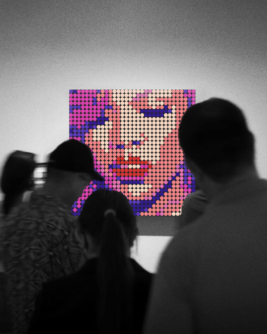 Varias personas observan una obra de arte en una galería. La pieza central es un retrato colorido hecho con piezas circulares tipo LEGO, que representa la portada del álbum Loud de Rihanna. La obra destaca con tonos rosados, violetas y rojos, mientras que el resto de la imagen está en blanco y negro, creando un contraste visual que centra la atención en la figura pixelada de la cantante.